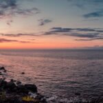 the sun is setting over the ocean on a rocky cliff