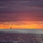 The sun is setting over the ocean with a lighthouse in the distance